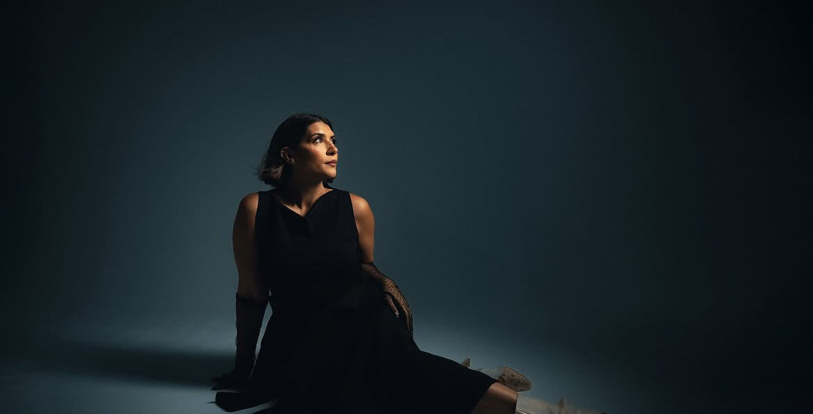 A woman in a black sleeveless dress sits on the floor against a dark blue-gray studio background, looking upward and to the side.